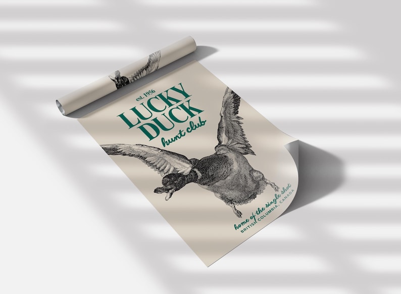 Puede incluir: Un p&oacute;ster enrollado con una detallada ilustraci&oacute;n en blanco y negro de un pato en vuelo. El p&oacute;ster tiene el texto "LUCKY DUCK hunt club" en verde azulado, con "est. 1956" arriba y "home of the single shot, BRITISH COLUMBIA, CANADA" abajo.