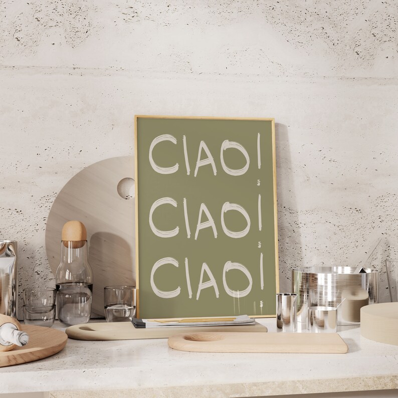 May include: A framed green poster with the word "CIAO" written three times in white. The poster is sitting on a white countertop with wooden chopping boards and other kitchen items.
