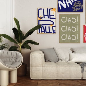 May include: A white sofa with three cushions, a potted banana plant, and three framed prints on the wall. The prints feature the words "Che Valle", "Ciao! Ciao! Ciao!" and "Na".