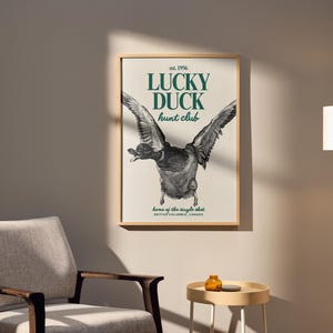Puede incluir: Un cuadro enmarcado con una ilustraci&oacute;n detallada de un pato en vuelo, con las alas extendidas. El texto "LUCKY DUCK hunt club" est&aacute; en verde, con "est. 1956" arriba y detalles de ubicaci&oacute;n abajo. La obra se muestra en una pared neutra.