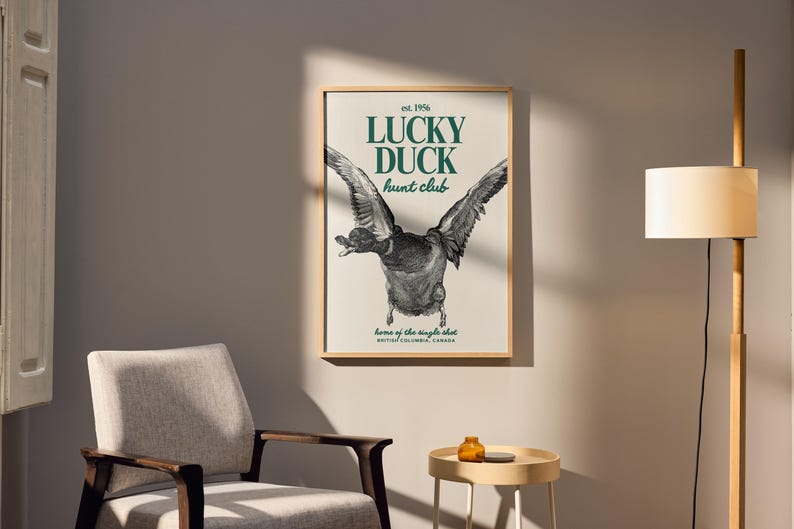 Puede incluir: Un cuadro enmarcado con una ilustraci&oacute;n detallada de un pato en vuelo, con las alas extendidas. El texto "LUCKY DUCK hunt club" est&aacute; en verde, con "est. 1956" arriba y detalles de ubicaci&oacute;n abajo. La obra se muestra en una pared neutra.