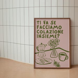 May include: A pink framed print with the text "Ti va se facciamo colazione insieme?" in green. The print features a green illustration of a coffee pot, a vase of flowers, a plate with cutlery, and a cup of coffee.