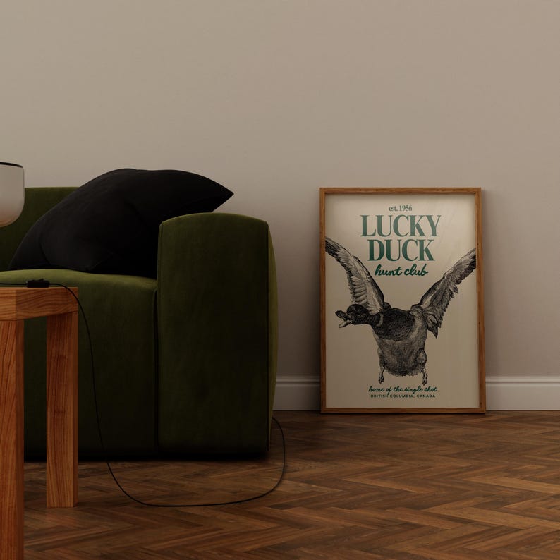 Puede incluir: Un cuadro enmarcado con el texto "LUCKY DUCK hunt club" y una ilustraci&oacute;n de un pato en vuelo. El cuadro est&aacute; en un marco de madera y se muestra cerca de un sof&aacute; verde y una mesa de madera.