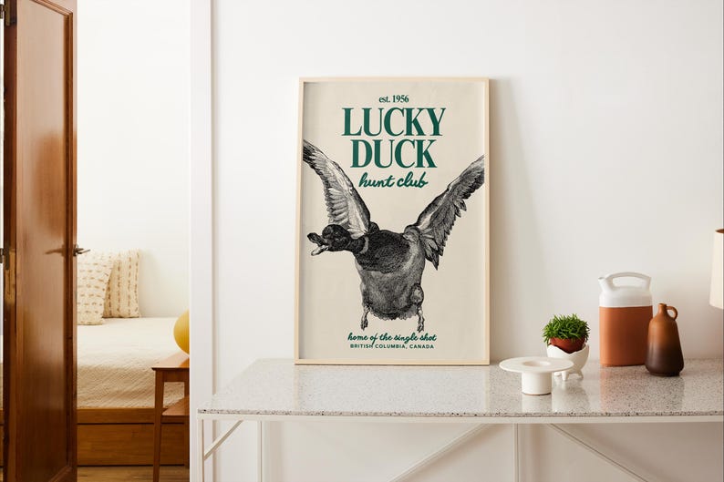 Puede incluir: Un p&oacute;ster enmarcado con una ilustraci&oacute;n detallada de un pato en vuelo. El texto verde dice "LUCKY DUCK hunt club", con "est. 1956" arriba y detalles de ubicaci&oacute;n abajo. Se muestra sobre una superficie blanca.