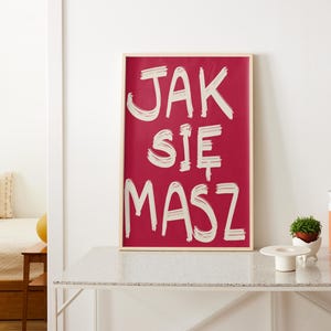 May include: A framed art print with the Polish phrase "JAK SIE MASZ" in white brushstrokes on a red background. The artwork is displayed on a white console table, with decorative items nearby.
