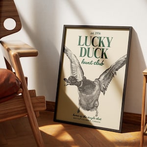 Puede incluir: L&aacute;mina enmarcada con una ilustraci&oacute;n de un pato en vuelo. El texto "LUCKY DUCK hunt club" es verde, con "est. 1956" arriba y "home of the single shot, BRITISH COLUMBIA, CANADA" abajo, sobre un fondo crema.