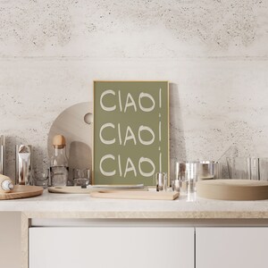 May include: A framed print with a green background and the word "CIAO" written three times in white. The print is sitting on a countertop with various kitchen items, including a wooden chopping board, a glass bottle, and a glass.