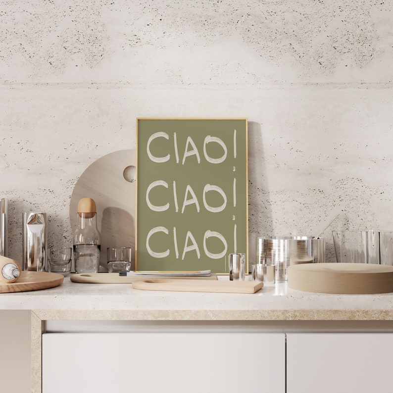 May include: A framed print with a green background and the word "CIAO" written three times in white. The print is sitting on a countertop with various kitchen items, including a wooden chopping board, a glass bottle, and a glass.