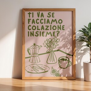 May include: A framed print with a pink background and green text that reads "Ti va se facciamo colazione insieme?"  The print features a green illustration of a coffee pot, a vase of flowers, a cup of coffee, and figs.