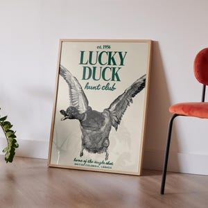 Puede incluir: P&oacute;ster enmarcado con una ilustraci&oacute;n detallada de un pato en vuelo. El texto "LUCKY DUCK hunt club" y "est. 1956" es visible. El p&oacute;ster est&aacute; apoyado contra una pared blanca, junto a una silla roja.