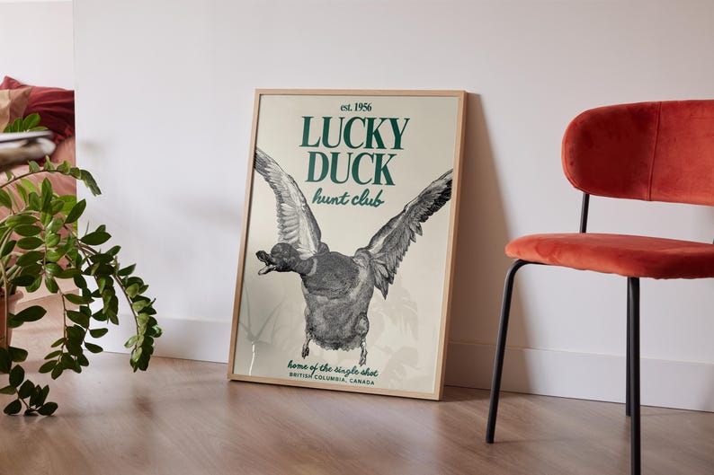 Puede incluir: P&oacute;ster enmarcado con una ilustraci&oacute;n detallada de un pato en vuelo. El texto "LUCKY DUCK hunt club" y "est. 1956" es visible. El p&oacute;ster est&aacute; apoyado contra una pared blanca, junto a una silla roja.