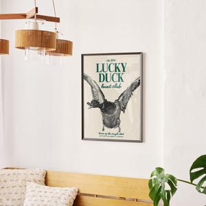 Puede incluir: Un p&oacute;ster enmarcado con el texto "LUCKY DUCK hunt club" y una ilustraci&oacute;n de un pato en vuelo. El p&oacute;ster est&aacute; en un marco negro y cuelga de una pared blanca. Tambi&eacute;n se ven una l&aacute;mpara de madera y una planta.