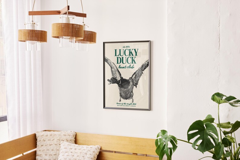 Puede incluir: Un p&oacute;ster enmarcado con el texto "LUCKY DUCK hunt club" y una ilustraci&oacute;n de un pato en vuelo. El p&oacute;ster est&aacute; en un marco negro y cuelga de una pared blanca. Tambi&eacute;n se ven una l&aacute;mpara de madera y una planta.