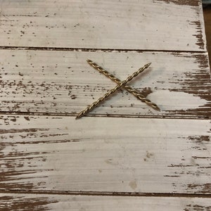 Peut inclure: Deux pi&egrave;ces de m&eacute;tal torsad&eacute;es dor&eacute;es formant un X sur une surface en bois blanc.