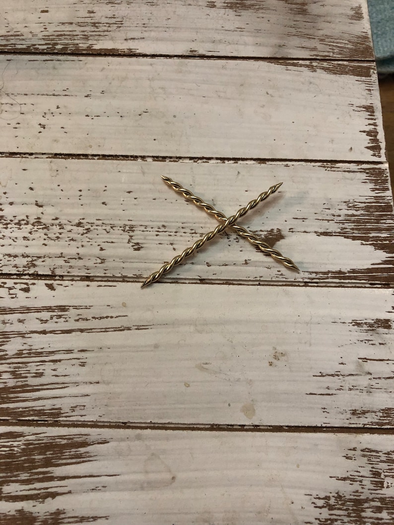 Peut inclure: Deux pi&egrave;ces de m&eacute;tal torsad&eacute;es dor&eacute;es formant un X sur une surface en bois blanc.