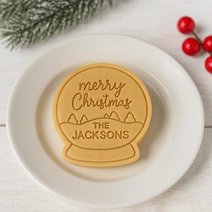 以下が含まれることがあります： 「merry Christmas」と「THE JACKSONS」の文字が刻まれた、スノードーム型の薄茶色のクッキー。白いお皿の上に置かれ、背景には常緑樹の枝と赤い実が添えられています。