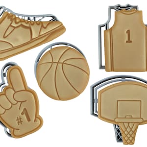 May include: A collection of five tan-colored cookie cutters in the shape of basketball-themed items. The set includes a basketball shoe, a basketball, a jersey with the number 1, a foam finger with the number 1, and a basketball hoop.