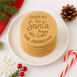 Peut inclure: Une pile de biscuits ronds et brun doré sur une assiette blanche. Les biscuits sont gravés des mots "cookies for Santa from THE JOHNSONS". La décoration de vacances comprend une pomme de pin, un sucre d'orge et du houx.