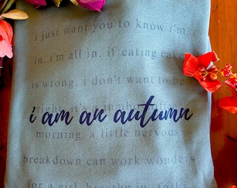 Könnte beinhalten: Ein teal-farbenes Sweatshirt mit dem Schriftzug "i am an autumn" in dunkelblauer Schrift. Zusätzlicher Text ist in einem helleren Farbton gedruckt. Das Bild ist mit Herbstblumen in Rot, Rosa und Gelb verziert.