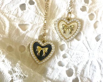 May include: Two gold necklaces with heart-shaped pendants. One pendant is white with a gold bow, the other is black with a gold bow. Both pendants are surrounded by a row of faux pearls.