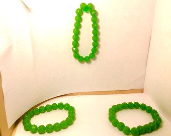 May include: Three green beaded bracelets. One bracelet is displayed vertically, while the other two are arranged horizontally. The beads have a faceted, rounded shape, and the bracelets are a vibrant, bright green color.