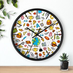 May include: A round wall clock with a black frame and a white face featuring a colorful pattern of school-themed illustrations including backpacks, trophies, globes, books, pencils, scissors, paper airplanes, and the letters A, B, and C.