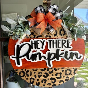 May include: Round wooden fall-themed door hanger with the text "Hey There Pumpkin". The top half is stained orange, the bottom half has a leopard print design. It is decorated with greenery and a bow with orange, black, and white accents.