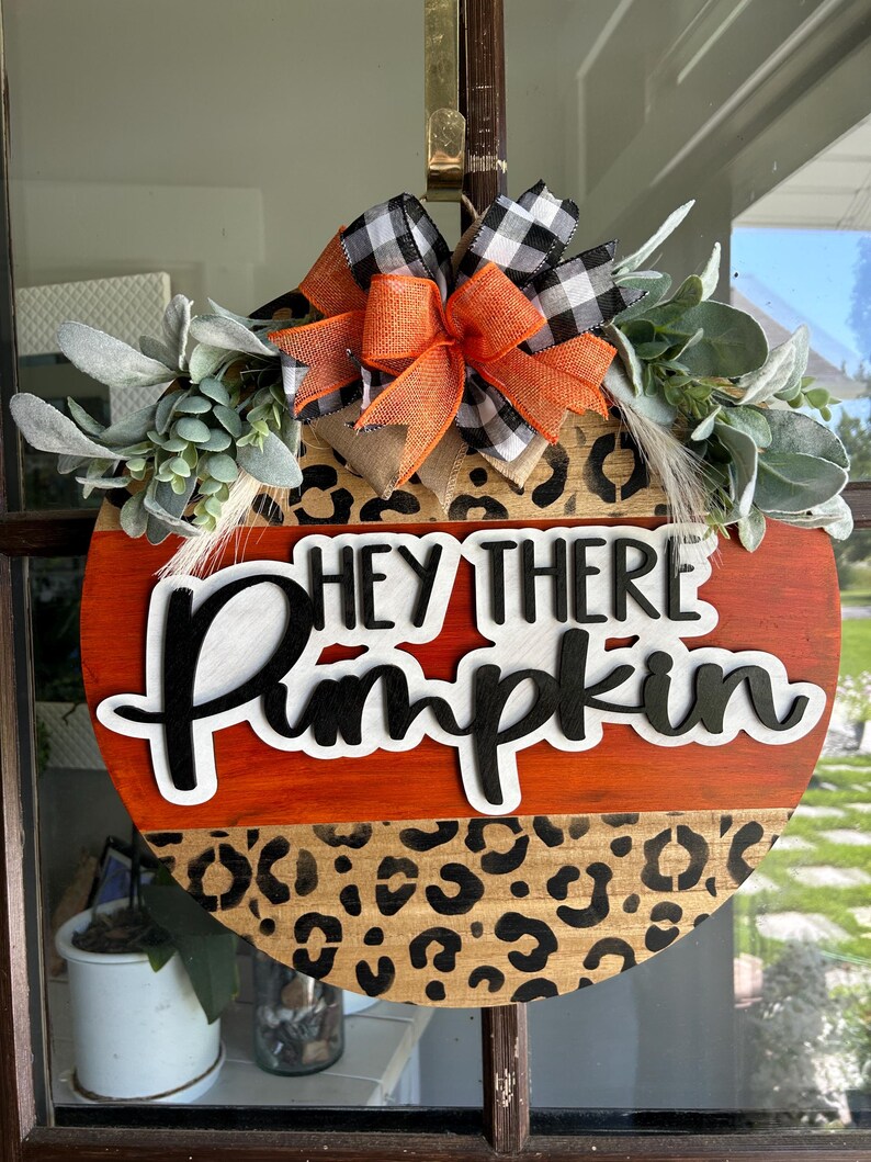 May include: Round wooden fall-themed door hanger with the text "Hey There Pumpkin". The top half is stained orange, the bottom half has a leopard print design. It is decorated with greenery and a bow with orange, black, and white accents.