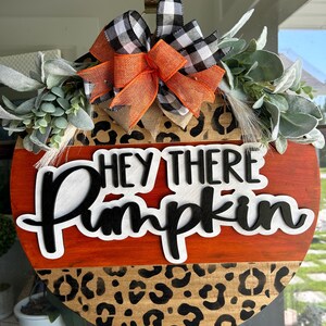 May include: Round wooden fall-themed sign with the text "HEY THERE Pumpkin". The sign features an orange and leopard print design, accented with a black and white buffalo check bow and greenery. A decorative piece for the autumn season.