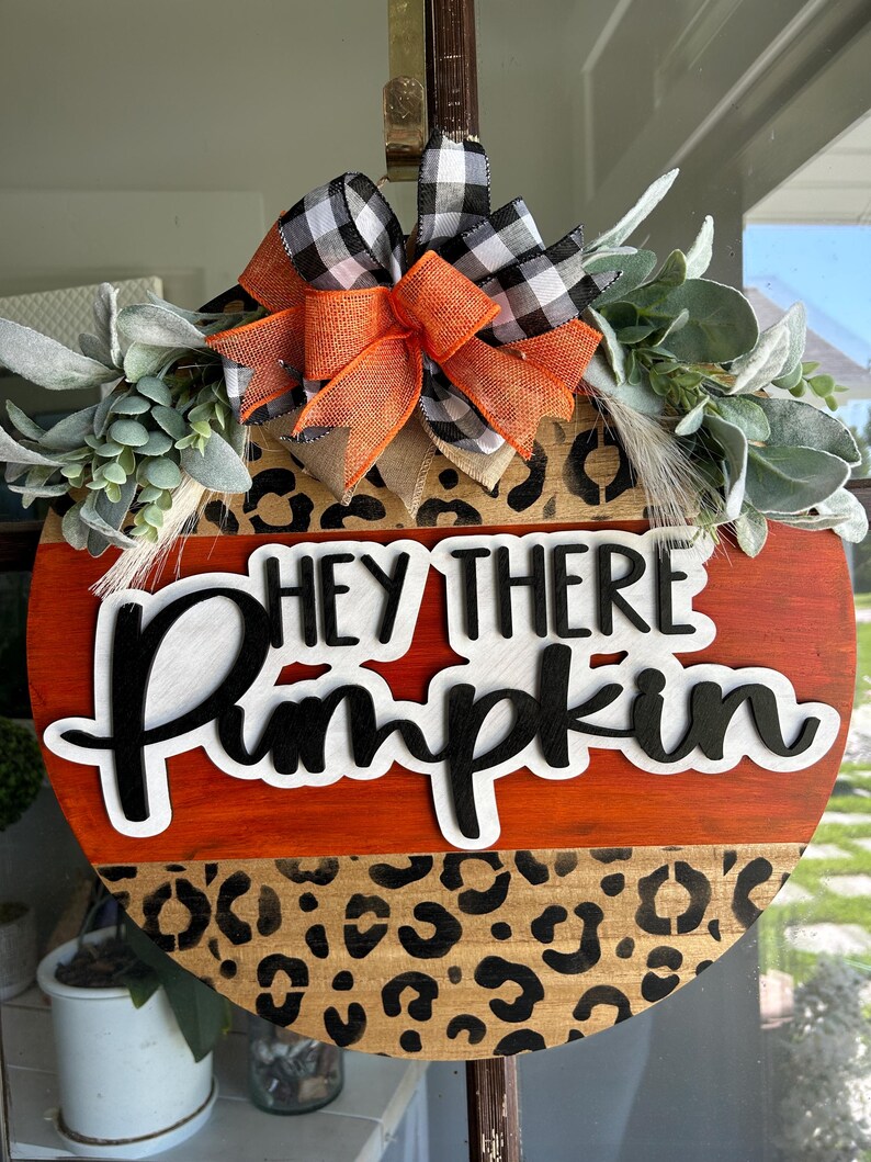 May include: Round wooden fall-themed sign with the text "HEY THERE Pumpkin". The sign features an orange and leopard print design, accented with a black and white buffalo check bow and greenery. A decorative piece for the autumn season.
