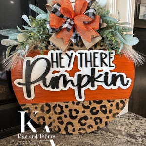May include: A round wooden pumpkin-shaped sign with the text "HEY THERE Pumpkin" in black and white. The top is orange, and the bottom has a leopard print design. It is decorated with greenery and a bow with orange, black, and white accents.