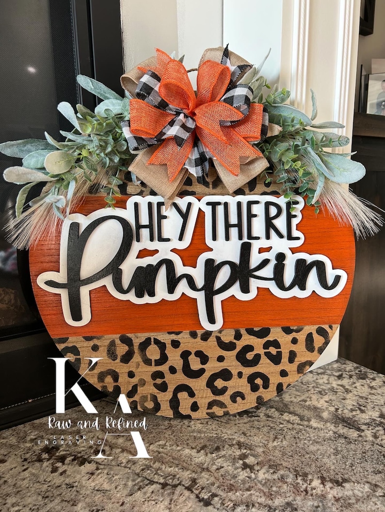 May include: A round wooden pumpkin-shaped sign with the text "HEY THERE Pumpkin" in black and white. The top is orange, and the bottom has a leopard print design. It is decorated with greenery and a bow with orange, black, and white accents.