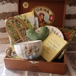 Peut inclure: Une boîte à cigares en bois Corina contenant une tasse blanche avec une illustration de Pierre Lapin, un petit livre jaune intitulé The World According to Mister Rogers, un cœur en feutrine verte brodé, une découpe de fenêtre en bois et un carré crocheté.