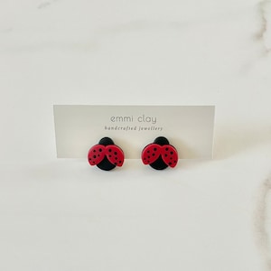 May include: A pair of red and black ladybug stud earrings. The earrings are handmade and feature a black background with red wings and black dots. The earrings are on a white surface with a card that says "emmi clay handcrafted jewellery".