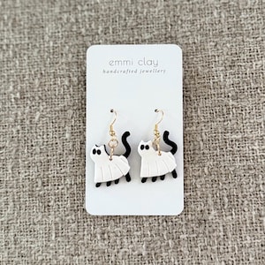 May include: Handmade cat earrings. The earrings are white with black details, including tails and feet, and are suspended from gold-coloured hooks. The earrings are presented on a white card with the text "emmi clay handcrafted jewellery".