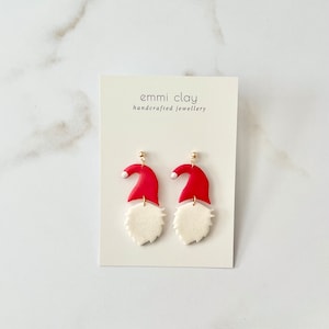May include: A pair of red and white gnome-themed earrings with gold hooks. The earrings are made of polymer clay and feature a red hat with a white pom-pom and a white beard.