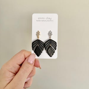 Peut inclure: Une paire de boucles d'oreilles en forme de feuille noire avec des accents dorés et argentés. Les boucles d'oreilles sont présentées sur une carte blanche avec le texte "emmi clay handcrafted jewellery".