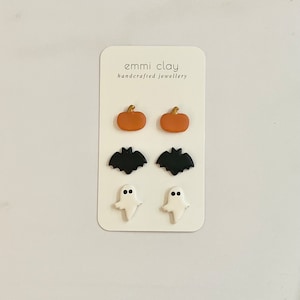 May include: A set of six Halloween-themed earrings, featuring a pair of orange pumpkins, a pair of black bats, and a pair of white ghosts. The earrings are displayed on a white card with the text "emmi clay handcrafted jewellery".