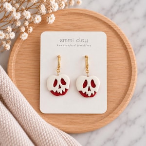 Peut inclure: Une paire de boucles d'oreilles créoles dorées avec des pendentifs en forme de pommes rouges recouvertes d'un glaçage blanc fondant, évoquant un crâne. Elles sont présentées sur une carte blanche marquée emmi clay handcrafted jewellery.