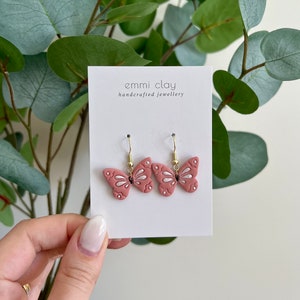 May include: A pair of pink butterfly earrings with silver accents and gold hooks. The earrings are displayed on a white card with the text "emmi clay handcrafted jewellery".