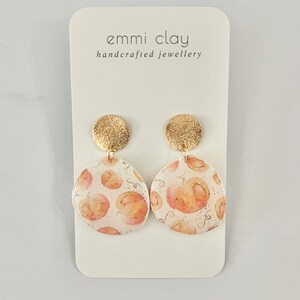 May include: A pair of gold and white pumpkin earrings. The earrings are made of polymer clay and feature a watercolour pumpkin design. The earrings are handmade and are perfect for autumn.
