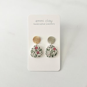 Peut inclure: Une paire de boucles d'oreilles faites à la main avec un disque en céramique blanche orné d'un motif floral vert et rouge et un dessus doré ou argenté. Les boucles d'oreilles sont présentées sur une carte blanche avec le texte "emmi clay handcrafted jewellery".