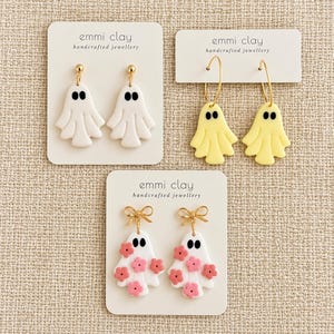 May include: Three pairs of earrings on white display cards labelled emmi clay handcrafted jewellery. One pair features white ghosts on gold studs, another has yellow ghosts on gold hoops, and the third shows white ghosts with pink flowers and gold bows.
