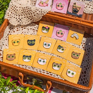 May include: A collection of 14 enamel pins featuring various cat designs. The pins are arranged in a wicker basket and feature different colors and patterns. The pins are all gold-toned and have the text "That's My Cat" on them.