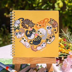 May include: A yellow spiral bound notebook with a heart-shaped design featuring multiple cartoon cats with different colored fur and large eyes. The notebook is on a small wooden easel and the text "@miibou.art" is at the bottom of the notebook.