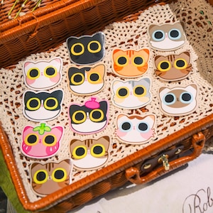 May include: A wicker basket filled with a variety of colorful cat stickers. The stickers feature different cat faces with various expressions and details, including a cat wearing a beret.