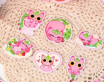 May include: A collection of pink strawberry cat stickers and a pink plush toy. The stickers feature a cat design with strawberry elements. One sticker is on a pink envelope. The stickers are on a cream-colored doily.