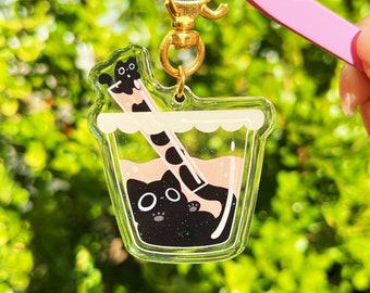 May include: A black cat charm with glitter inside a clear plastic cup with a black and white striped straw. The charm is attached to a gold metal key chain.