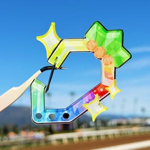 Puede incluir: Un amuleto geom&eacute;trico colorido con un degradado arco&iacute;ris, detalles de estrellas y una forma de gema verde. El amuleto es sostenido por unas pinzas contra un cielo azul y un hip&oacute;dromo.