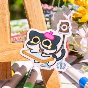 May include: Two black and white cartoon cat stickers with pink accents. The stickers feature a cat wearing a beret and holding a paintbrush, sitting at an easel. The stickers are on a white tablecloth with a floral pattern.